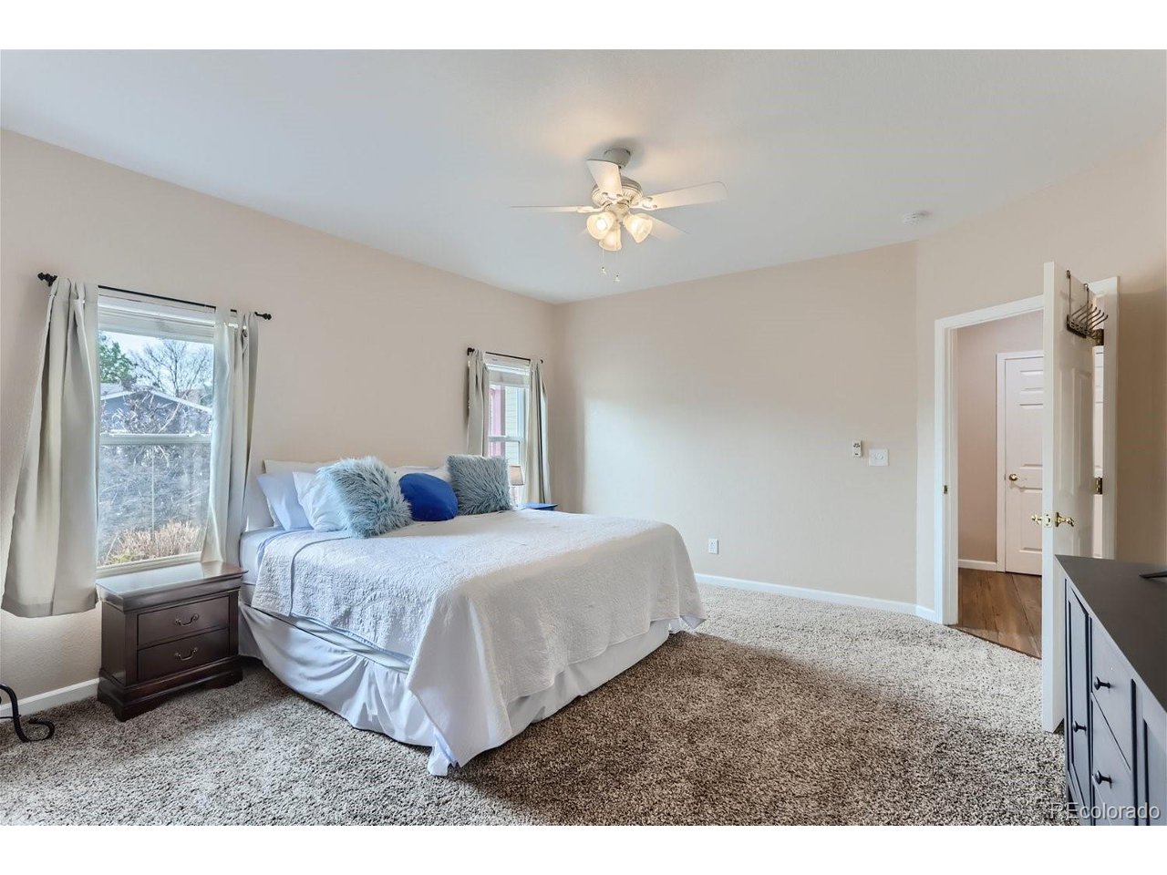 Bedroom with a white bed, fluffy accent pillows, two curtained windows, and beige carpet.