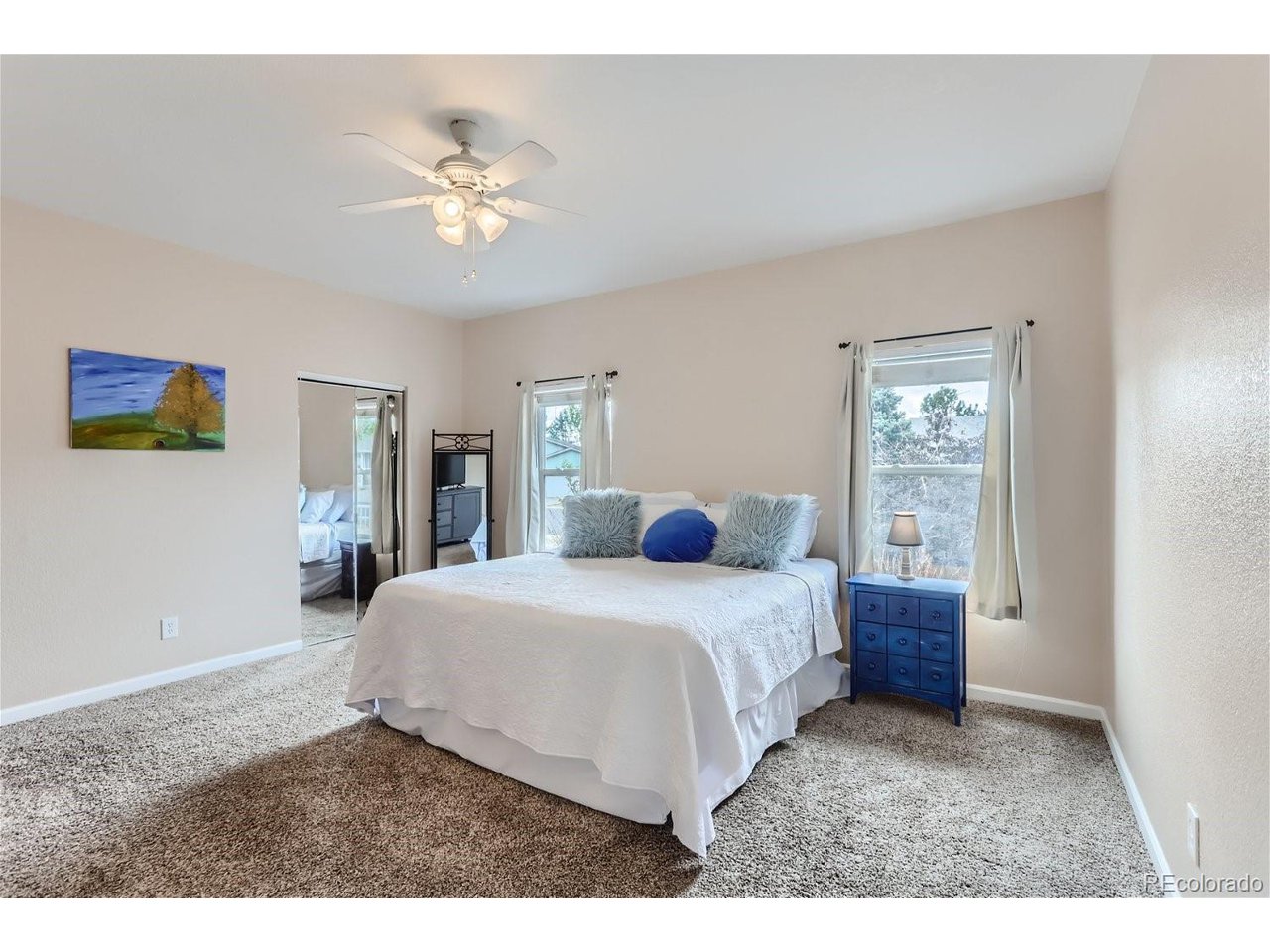 Bedroom with a white bed, blue bedside chest, mirrored closet door, and ceiling fan.