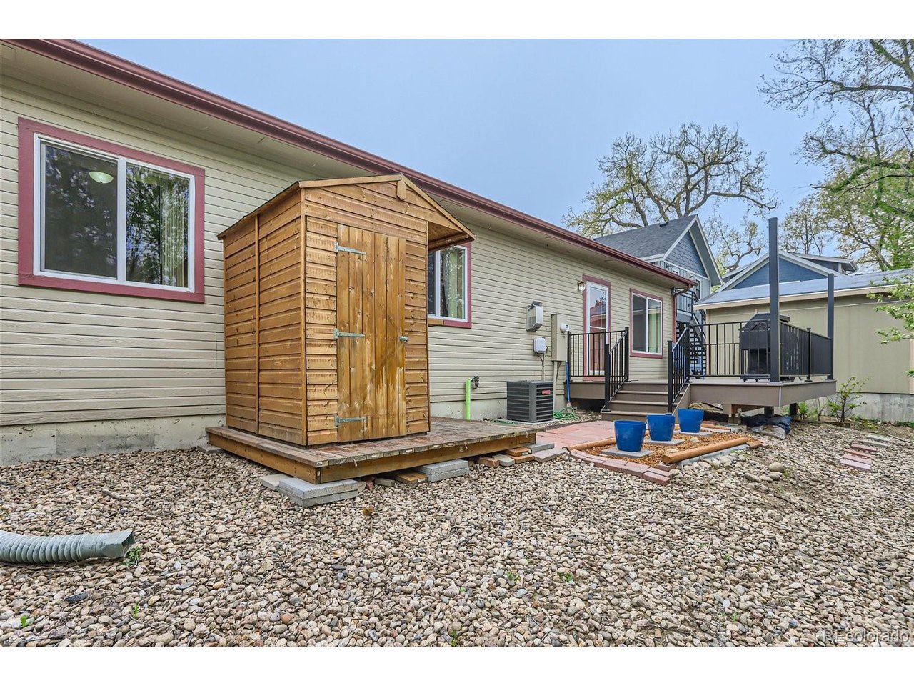 Rear of a beige-sided house with a wooden shed, raised deck, and gravel yard.