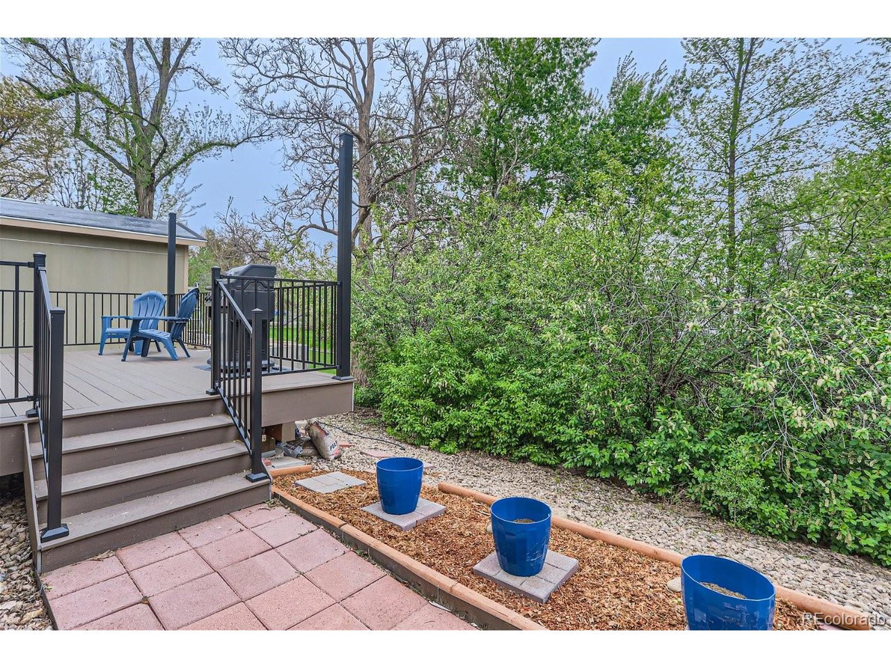 Raised backyard deck with black railings, blue chairs, and steps beside a paver patio and garden bed.