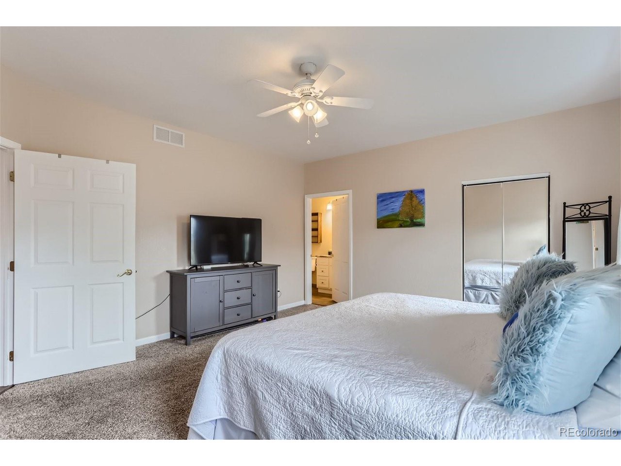 Furnished bedroom with a white bed, blue accent pillows, mirrored closet doors, and a ceiling fan.