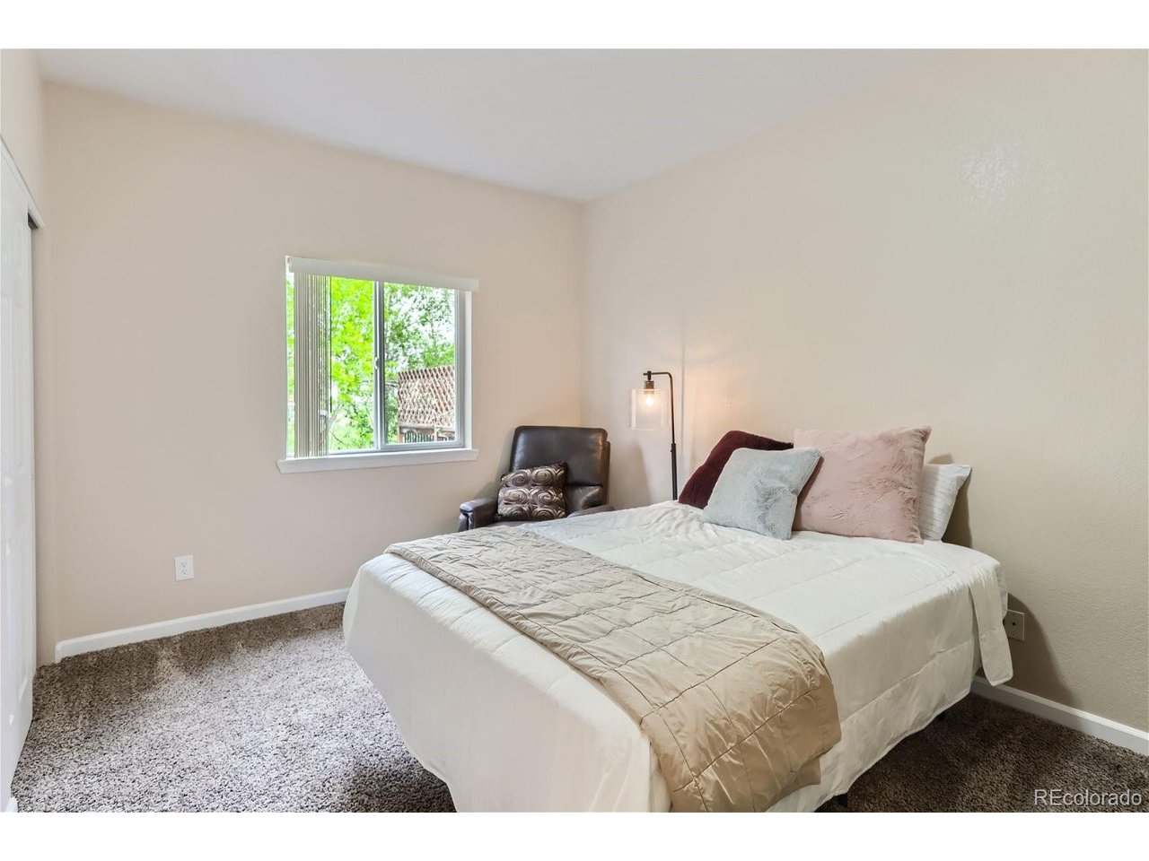 Staged bedroom with a white bed, pastel pillows, leather armchair, and window overlooking leafy trees.