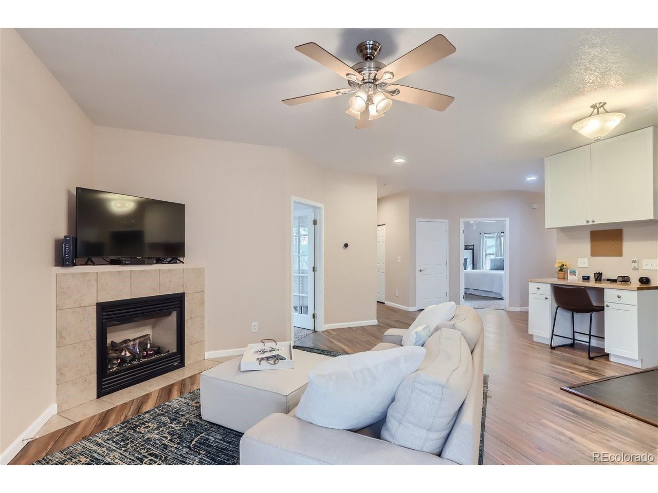 Living room with a tiled gas fireplace, wall-mounted TV, pale sofas, and an open kitchen.
