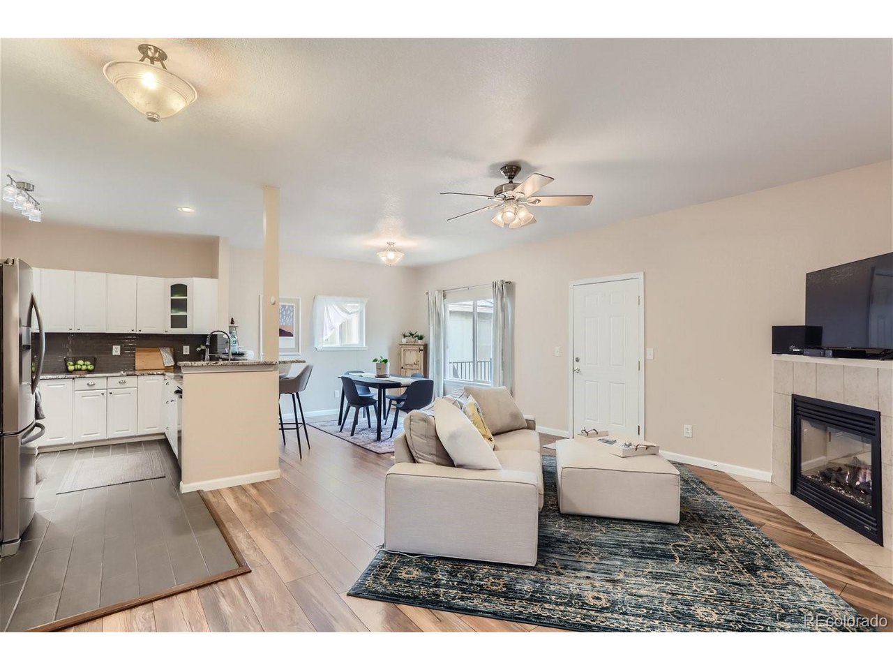 Open-plan living room with beige sectional, dark patterned rug, fireplace, and adjoining kitchen.