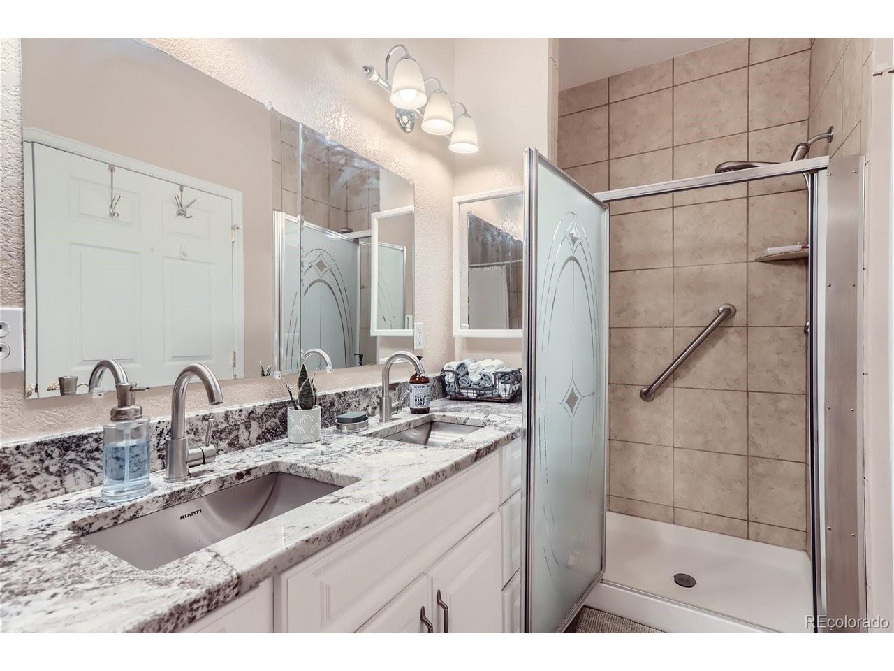 Bathroom with a double vanity and granite counters beside a tiled shower with a frosted glass door.