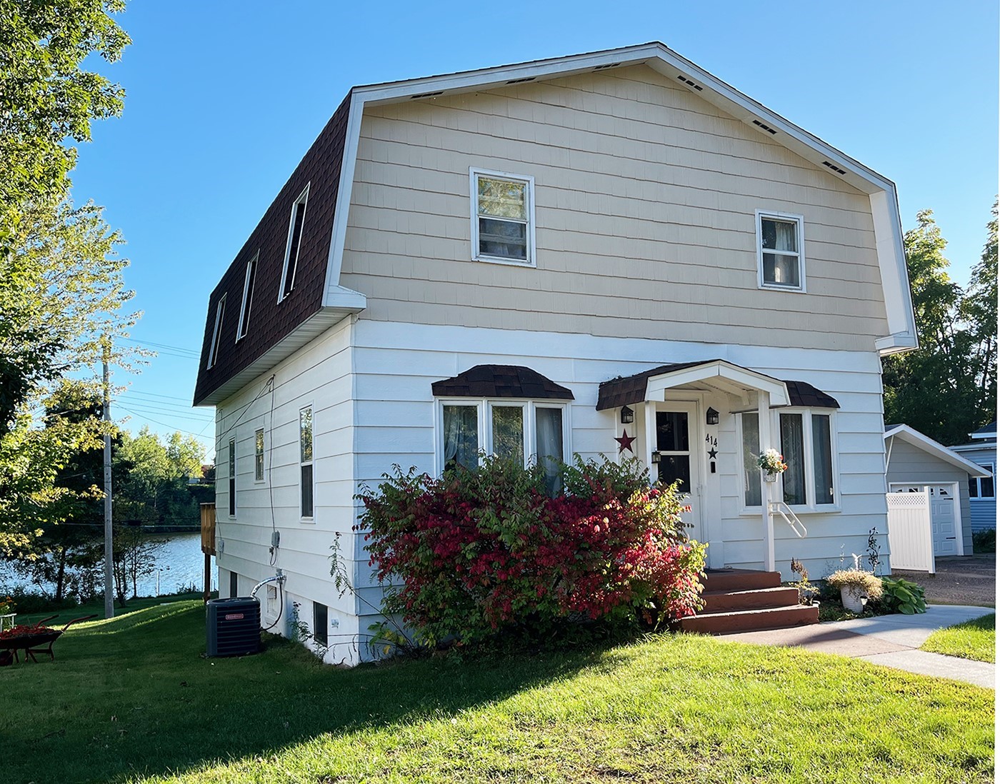 Two-story white house with dark gambrel roof, landscaped entry, and water visible beside it.