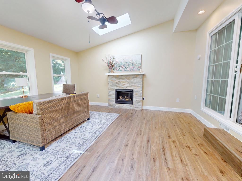 Living room with a stone fireplace, wicker seating, light wood floors, and large windows.