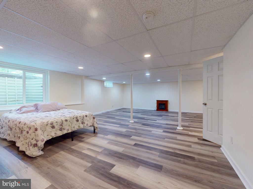 Finished basement with gray plank flooring, a bed by the window, support posts, and an electric fireplace.