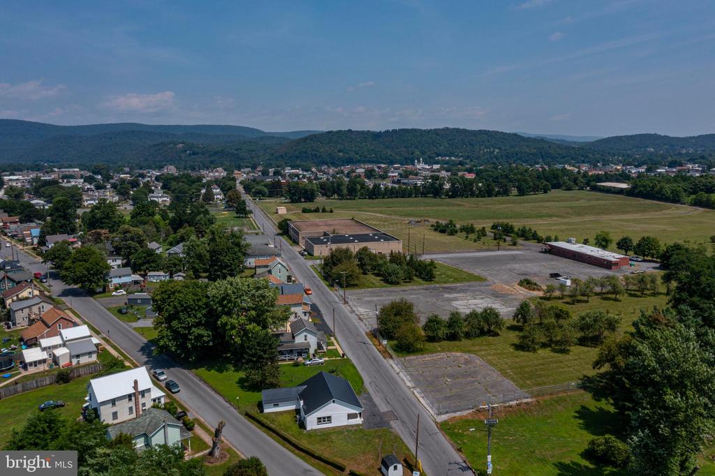 Aerial view of homes and open lots beside a road, with wooded hills beyond.