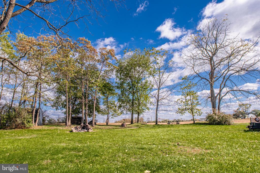 Grassy backyard with mature trees, a stone fire pit, and open fields beyond.