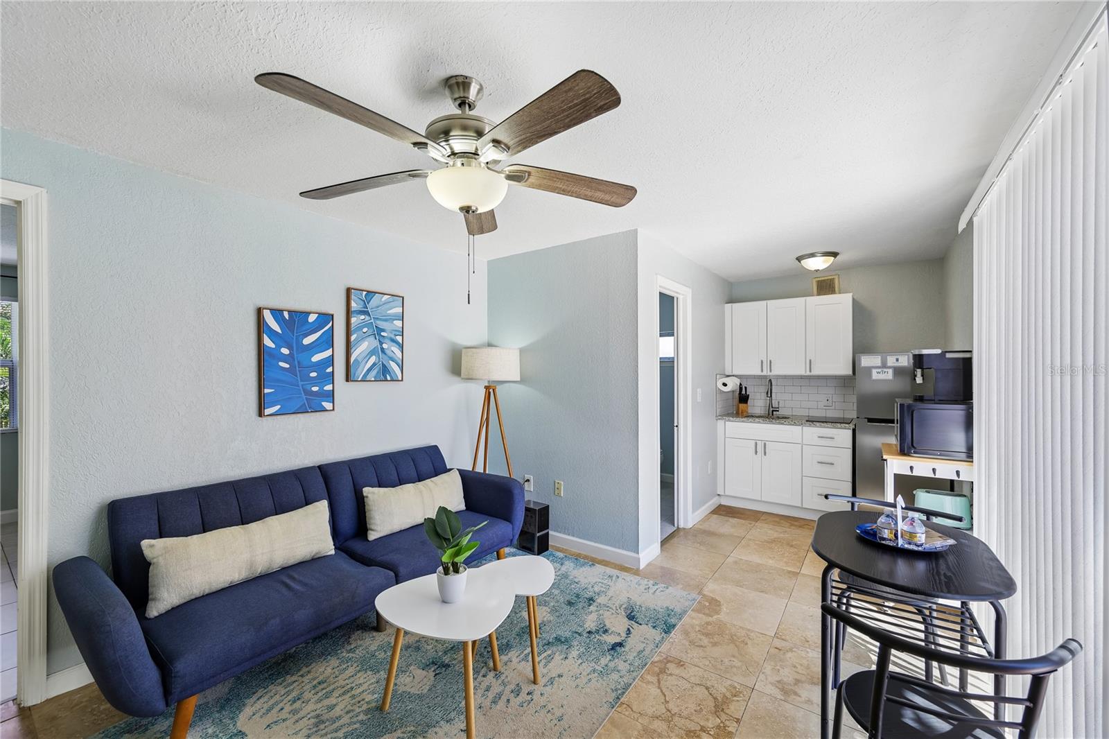 Living room with a navy sofa and ceiling fan beside a compact white-cabinet kitchenette.