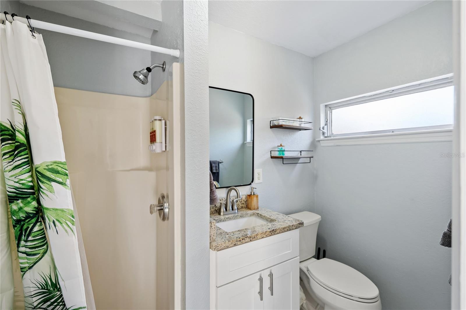 Bathroom with a leaf-print shower curtain, stone-counter vanity, toilet, and high horizontal window.