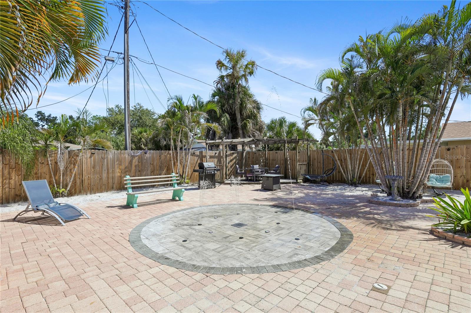 Fenced backyard patio with brick pavers, palm trees, pergola seating, and a grill.