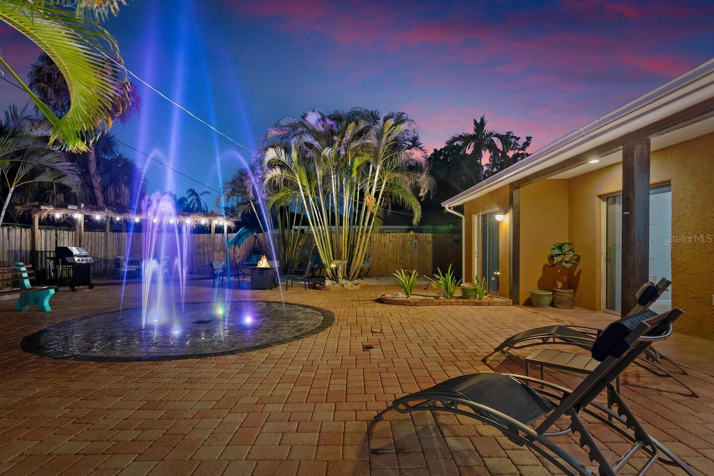 Backyard patio with a lit fountain pool, tropical palms, and lounge chairs at night.