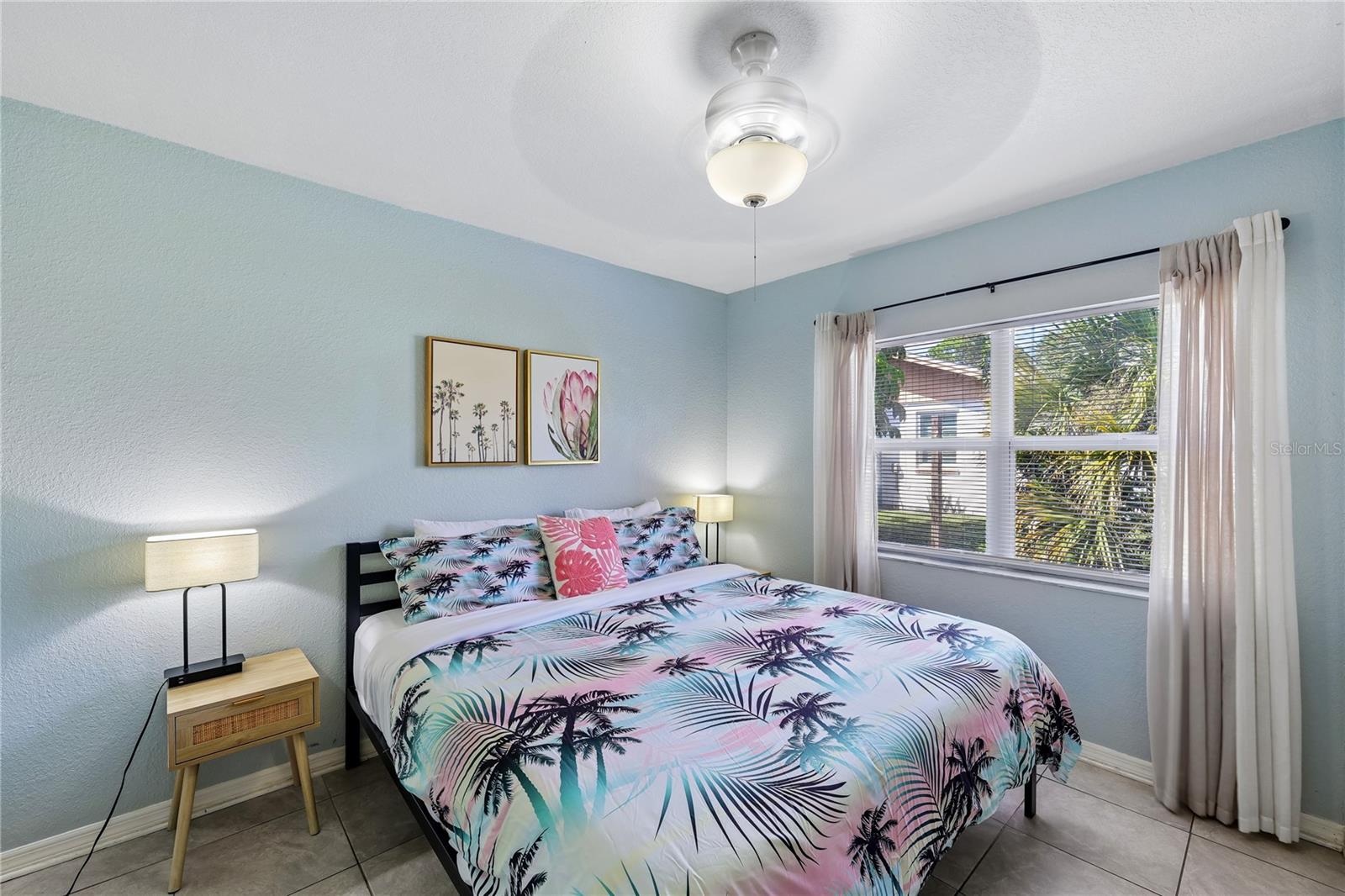 Bedroom with tropical palm-print bedding, pale blue walls, and a large curtained window.