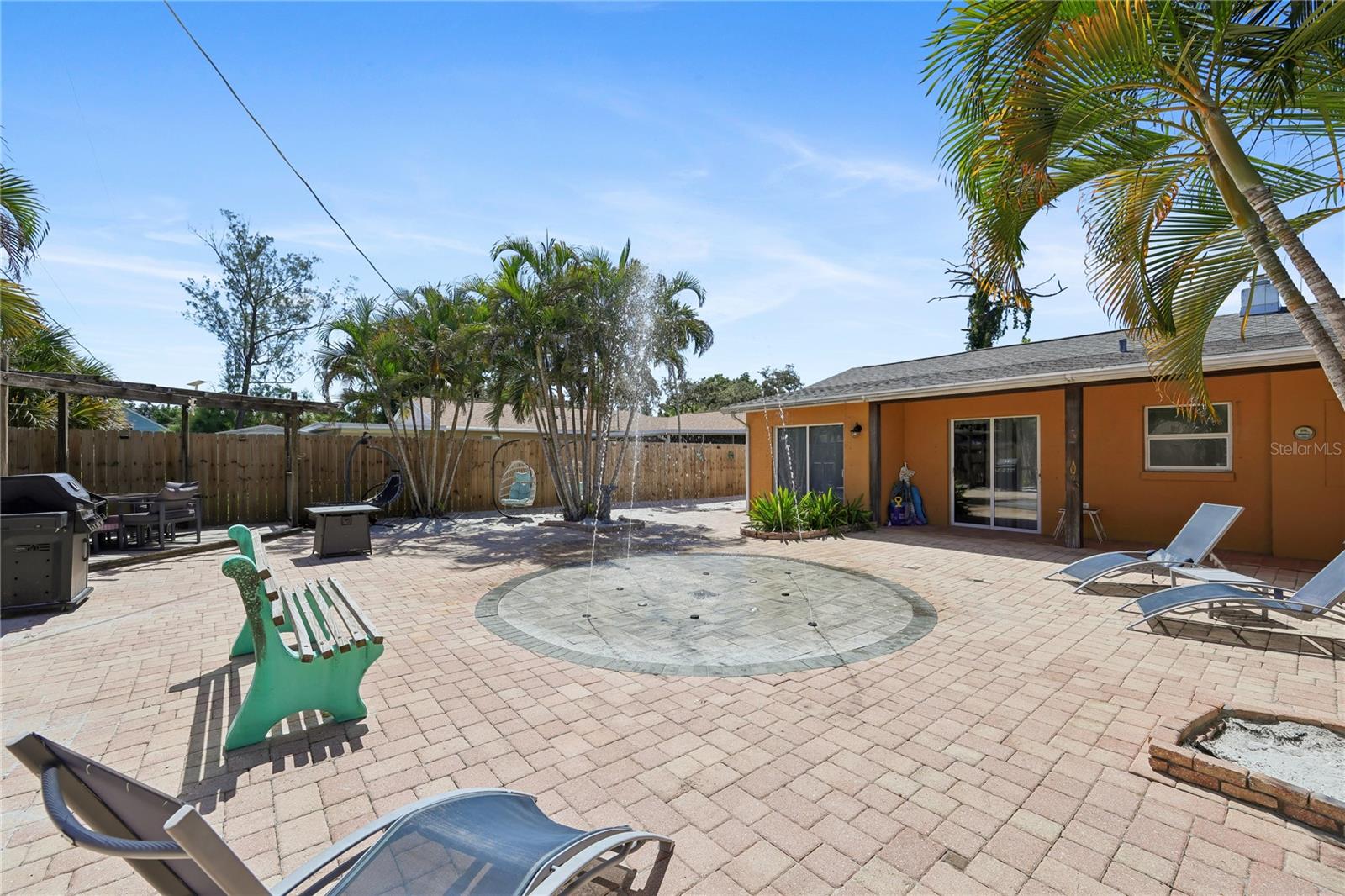 Backyard patio with a circular fountain, brick pavers, palm trees, and lounge chairs.
