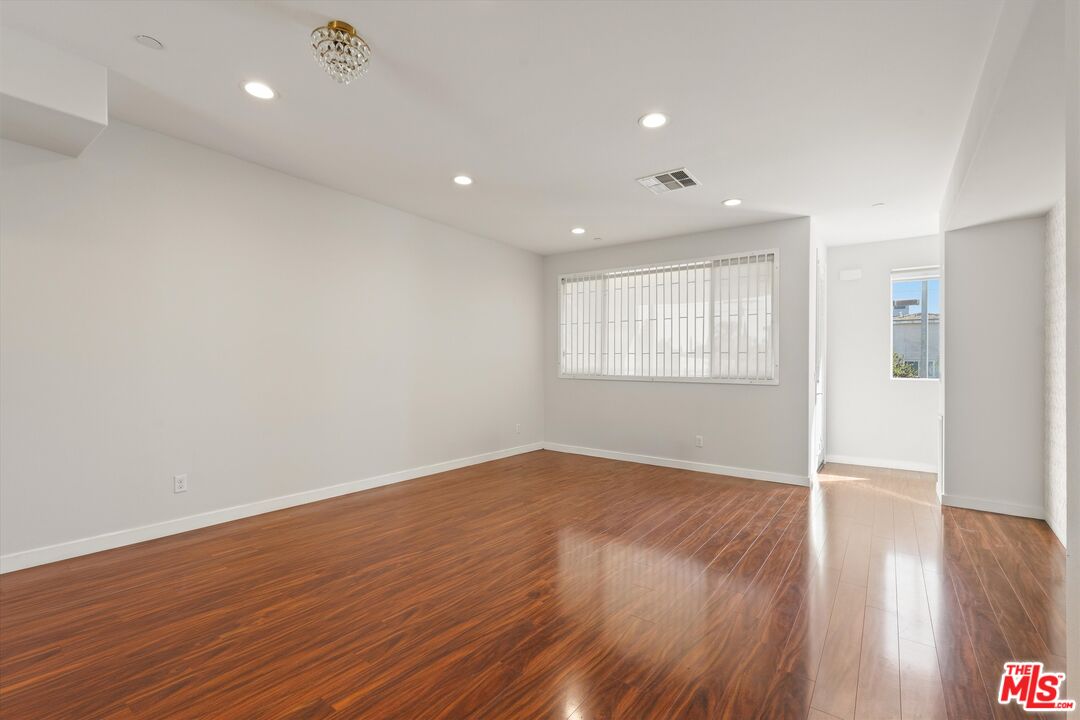 Vacant living room with polished wood floors, recessed lights, and a barred window.