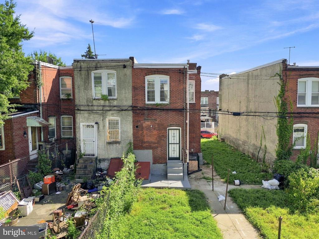Rear of a Philadelphia rowhouse with brick and stucco walls and a cluttered, overgrown backyard.