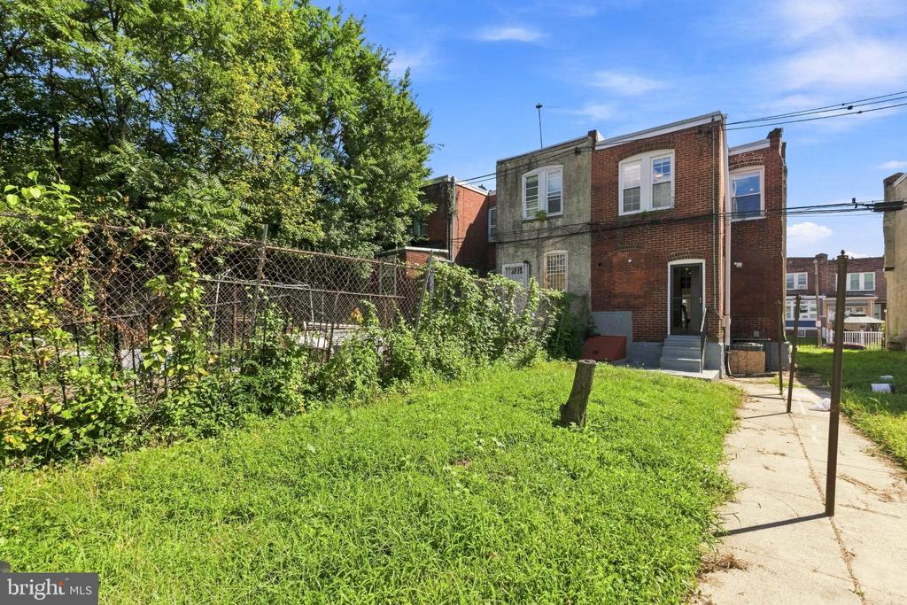 Rear of a brick rowhouse with a grassy yard, chain-link fence, and concrete walkway.