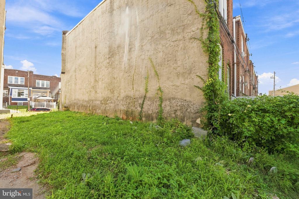 Overgrown vacant lot beside brick row homes, bordered by a tall weathered wall.