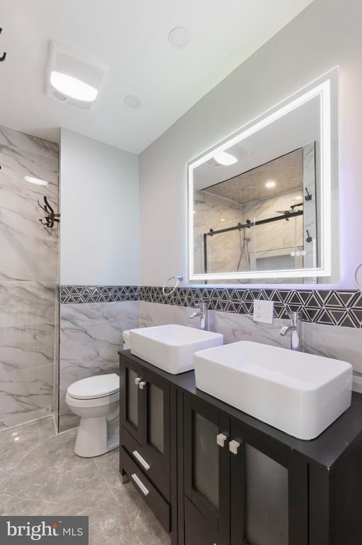 Bathroom with double vessel sinks, a dark vanity, marble-look tile, and a large illuminated mirror.