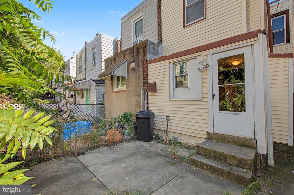 Rear of a beige-sided row house with a white back door opening onto a concrete patio and fenced yard.