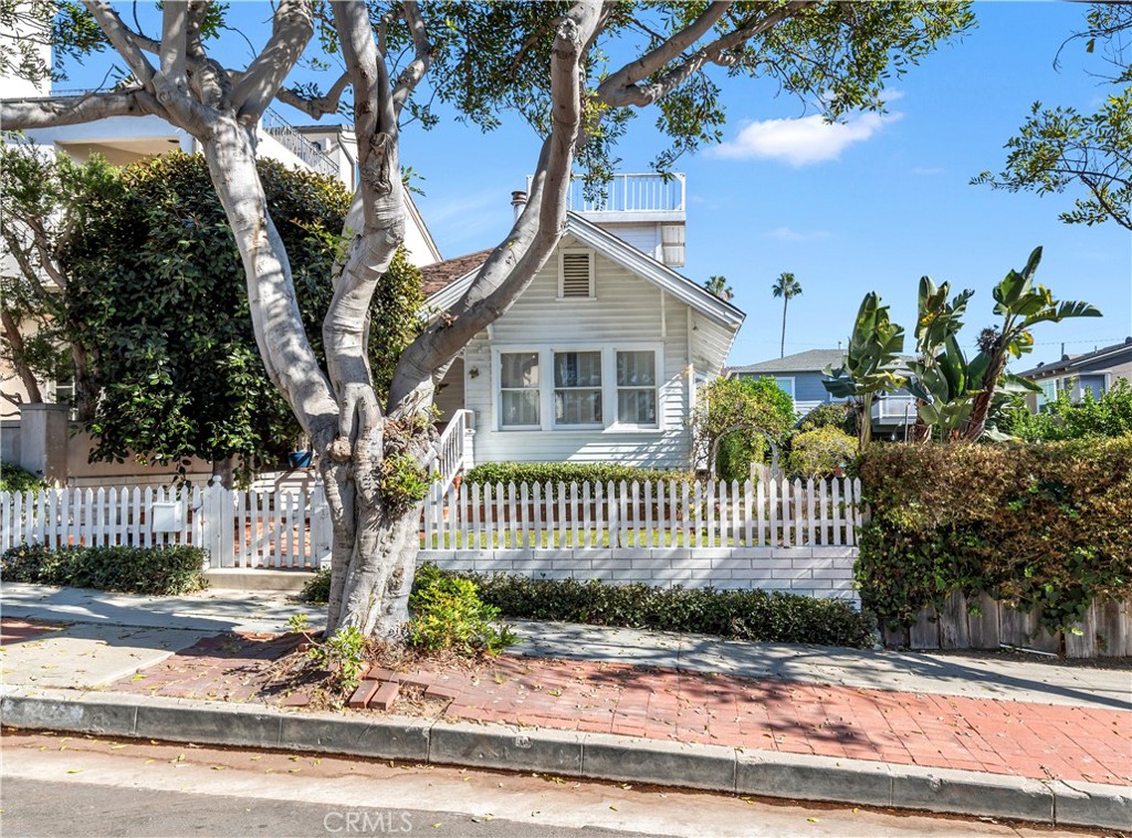 White coastal-style house behind a picket fence, shaded by a large tree.