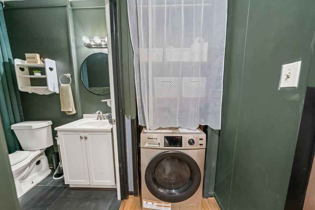 Bathroom with a white vanity and toilet beside a front-loading washer behind a curtain.