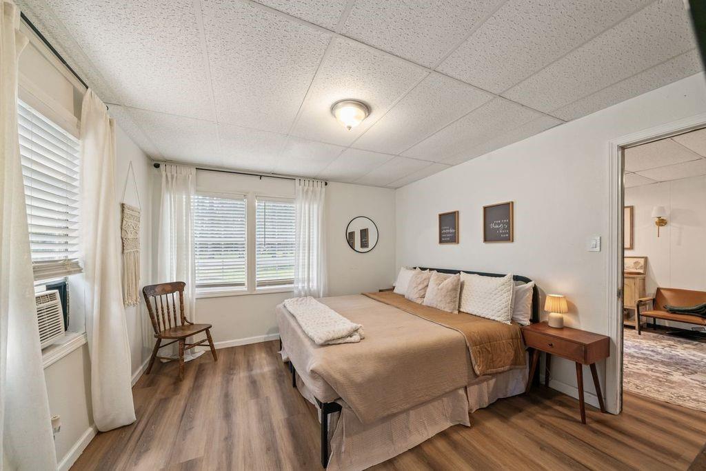 Bedroom with a neatly made bed, wood-look flooring, and windows with white curtains.