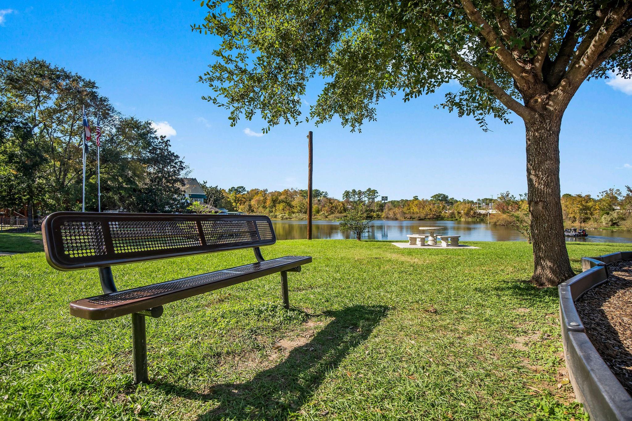 Lakeside community park with a metal bench, picnic tables, and mature trees.