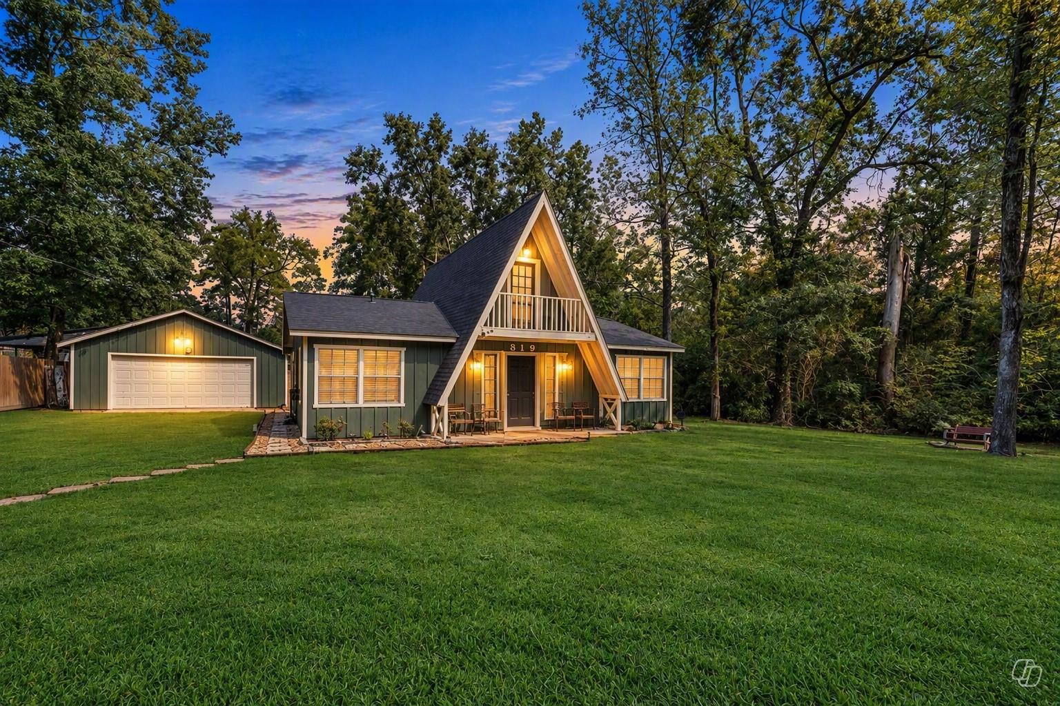 Green A-frame house with a detached garage, broad lawn, and wooded surroundings at dusk.
