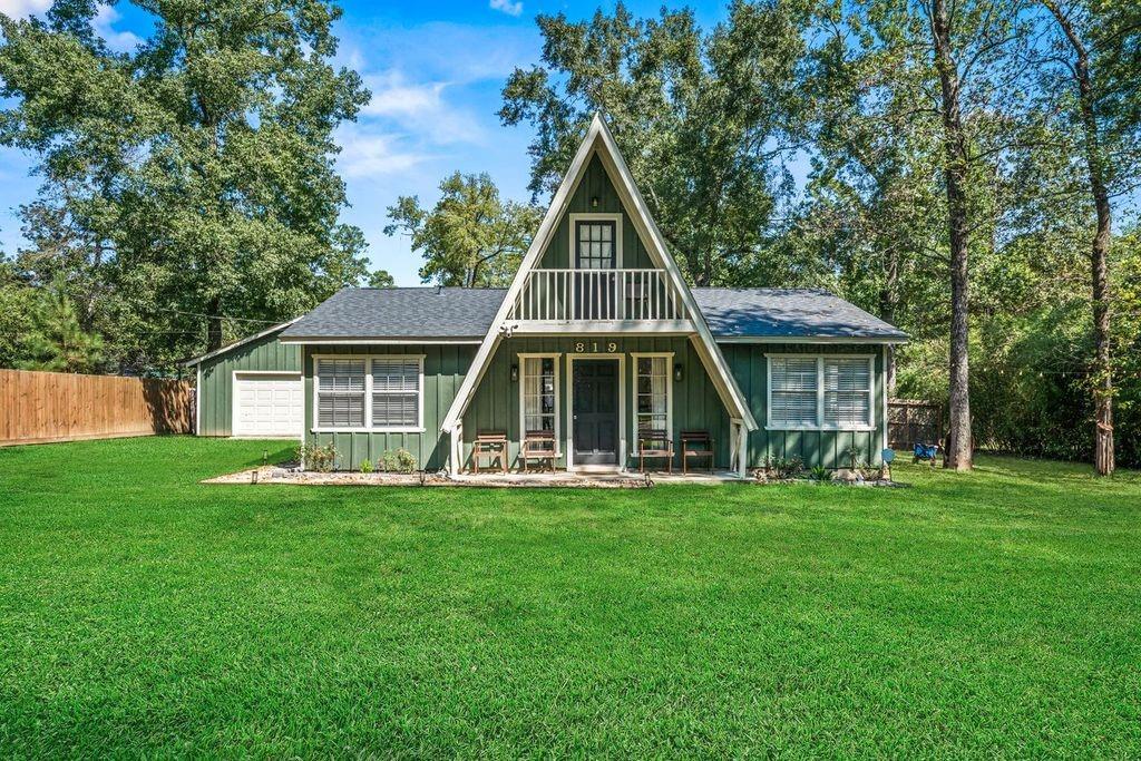 Green A-frame house with a small balcony, broad lawn, and mature trees.