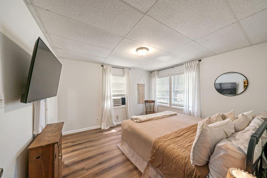 Bedroom with a large bed, wood-look flooring, and windows with white curtains.