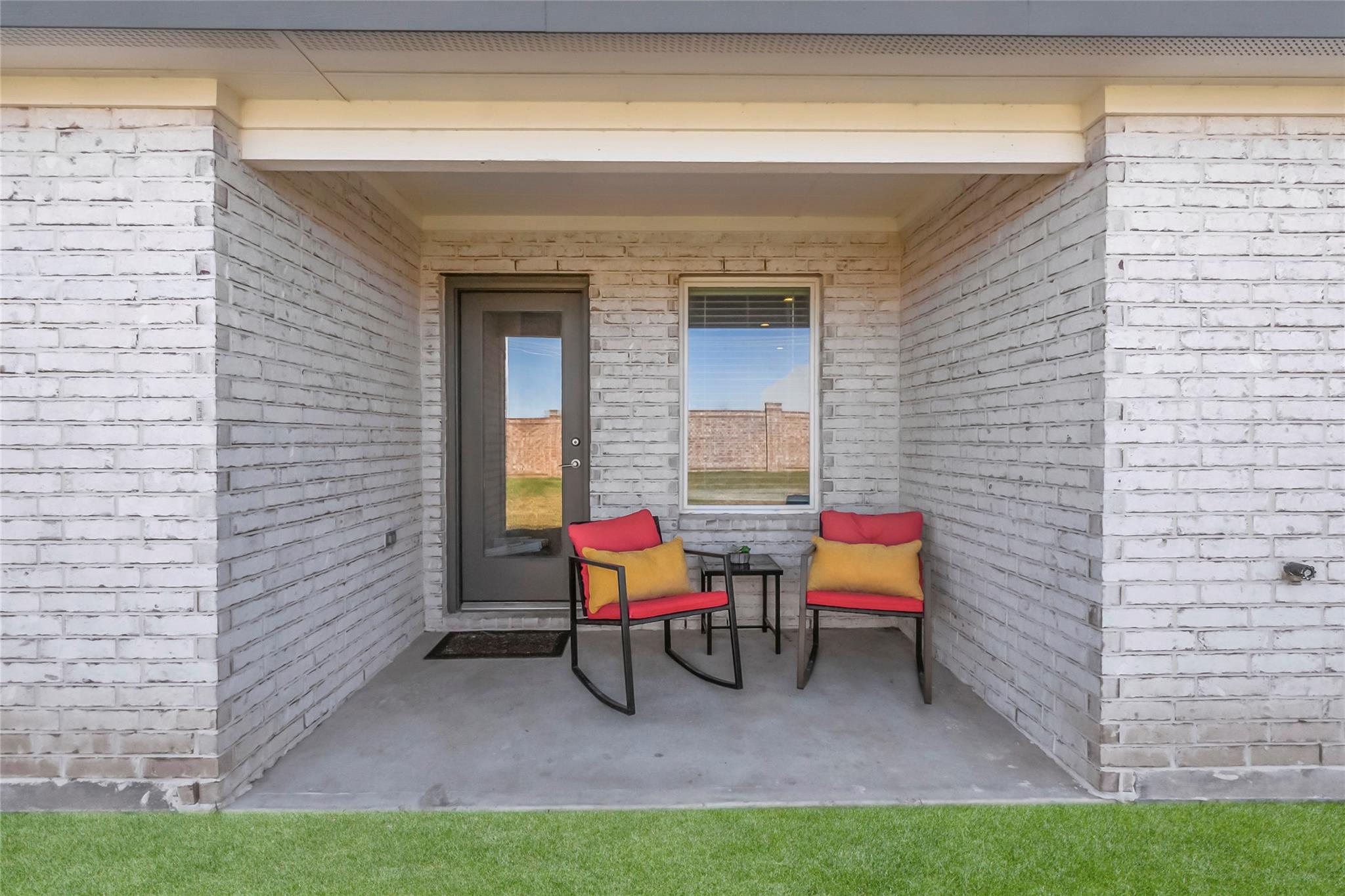 Covered white-brick front porch with two red chairs, yellow pillows, and a small table.
