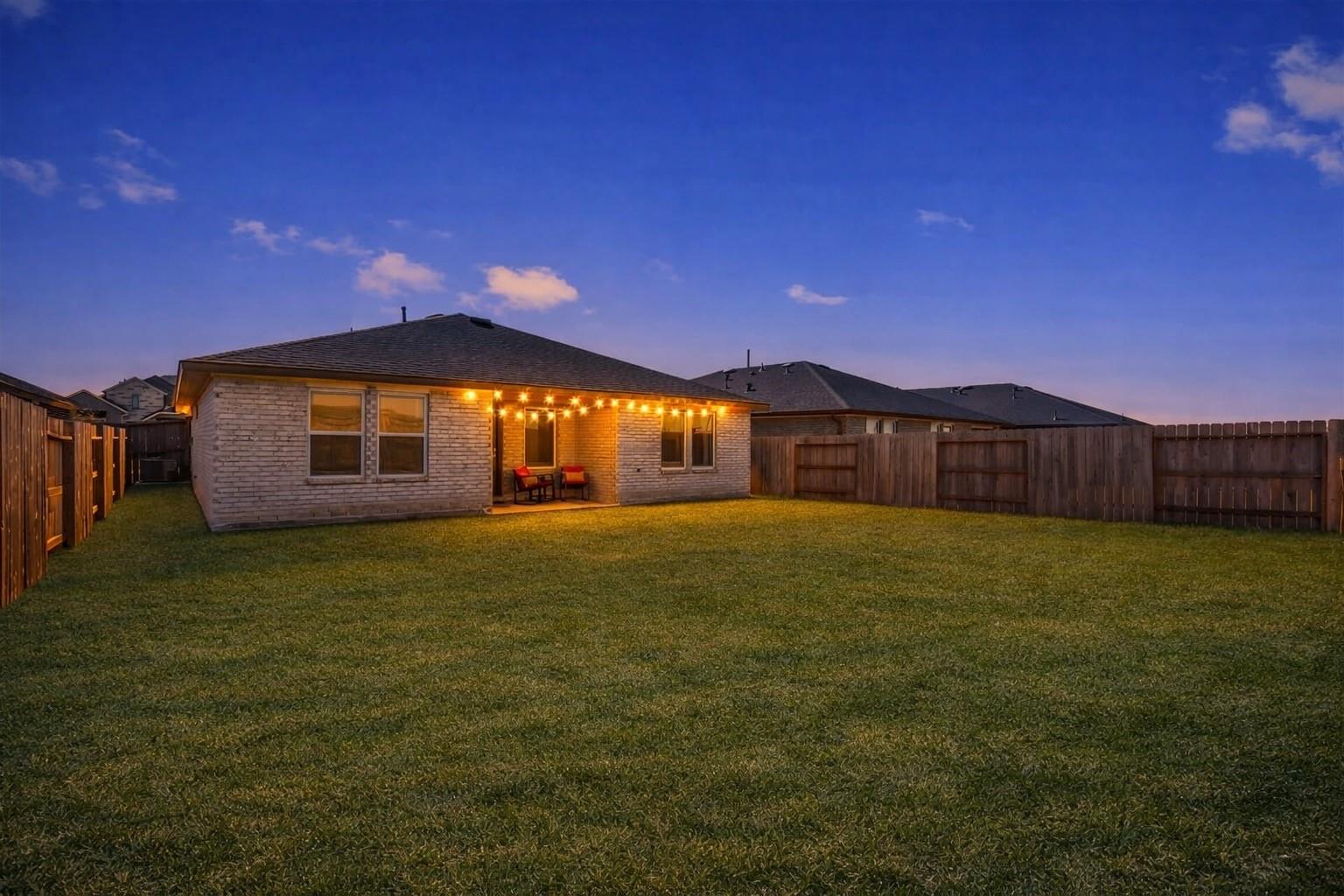 Brick home’s rear exterior at dusk, with a spacious fenced lawn and a string-lit covered patio.