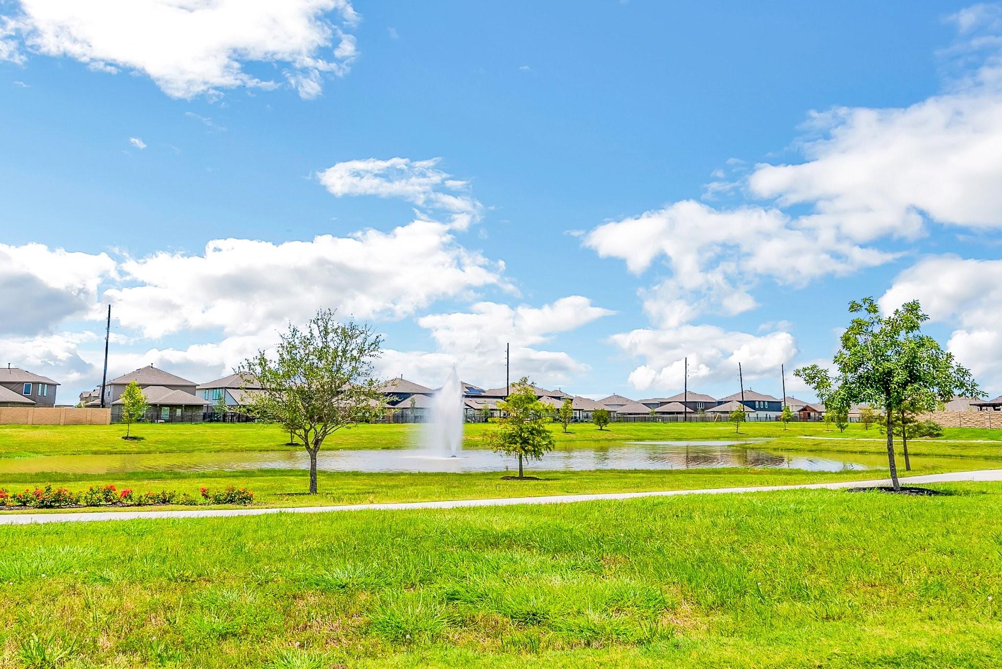 Community pond and fountain beside a grassy path, with homes in the background.