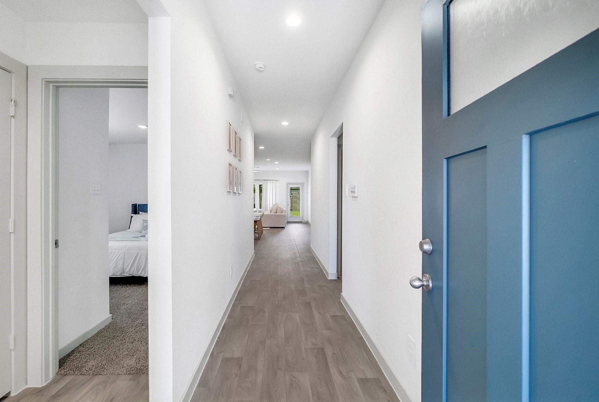 Bright hallway with gray wood-look floors and a blue paneled front door; bedroom and living area visible beyond.