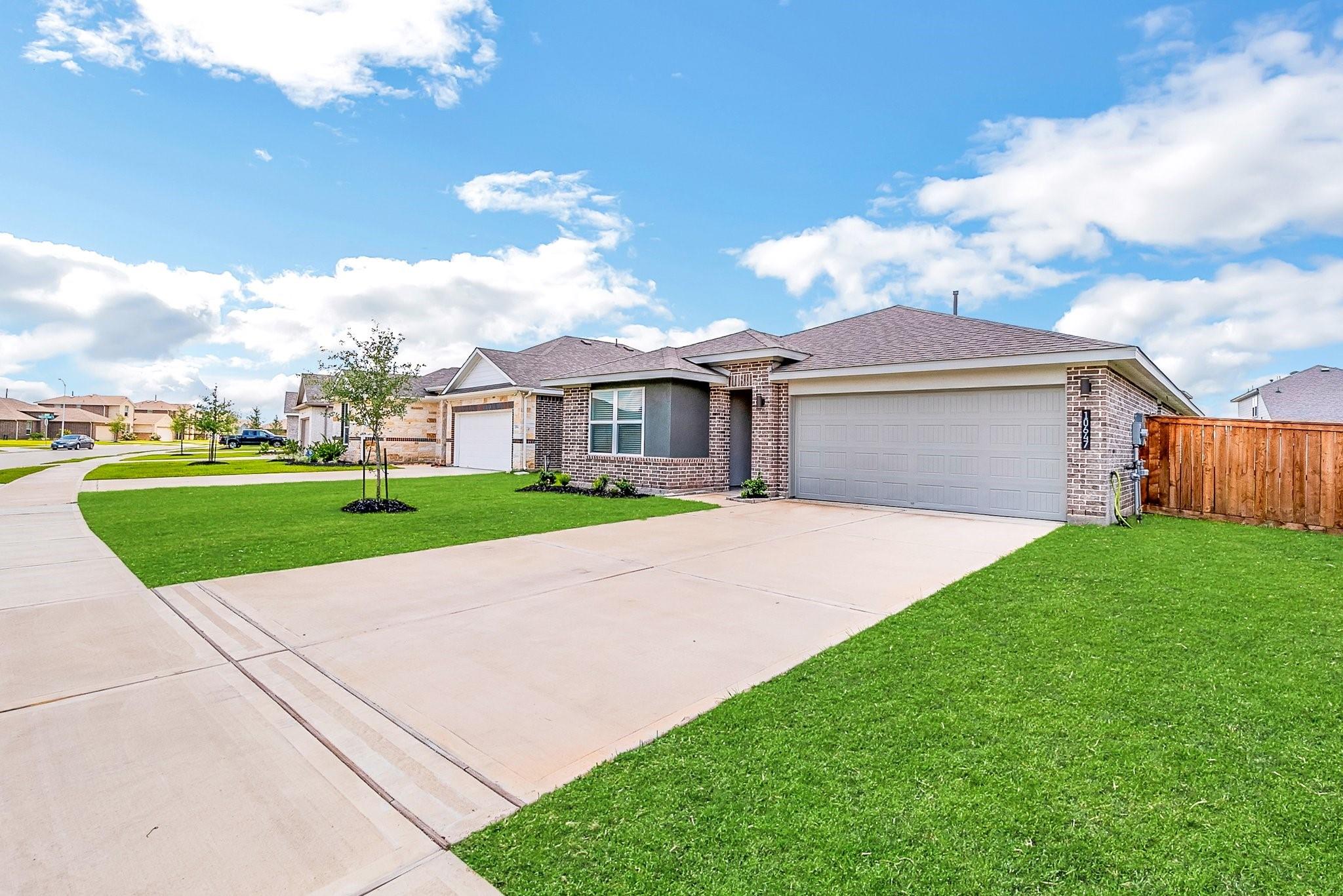 Brick single-story home with a wide garage, broad driveway, green lawn, and wooden side fence.