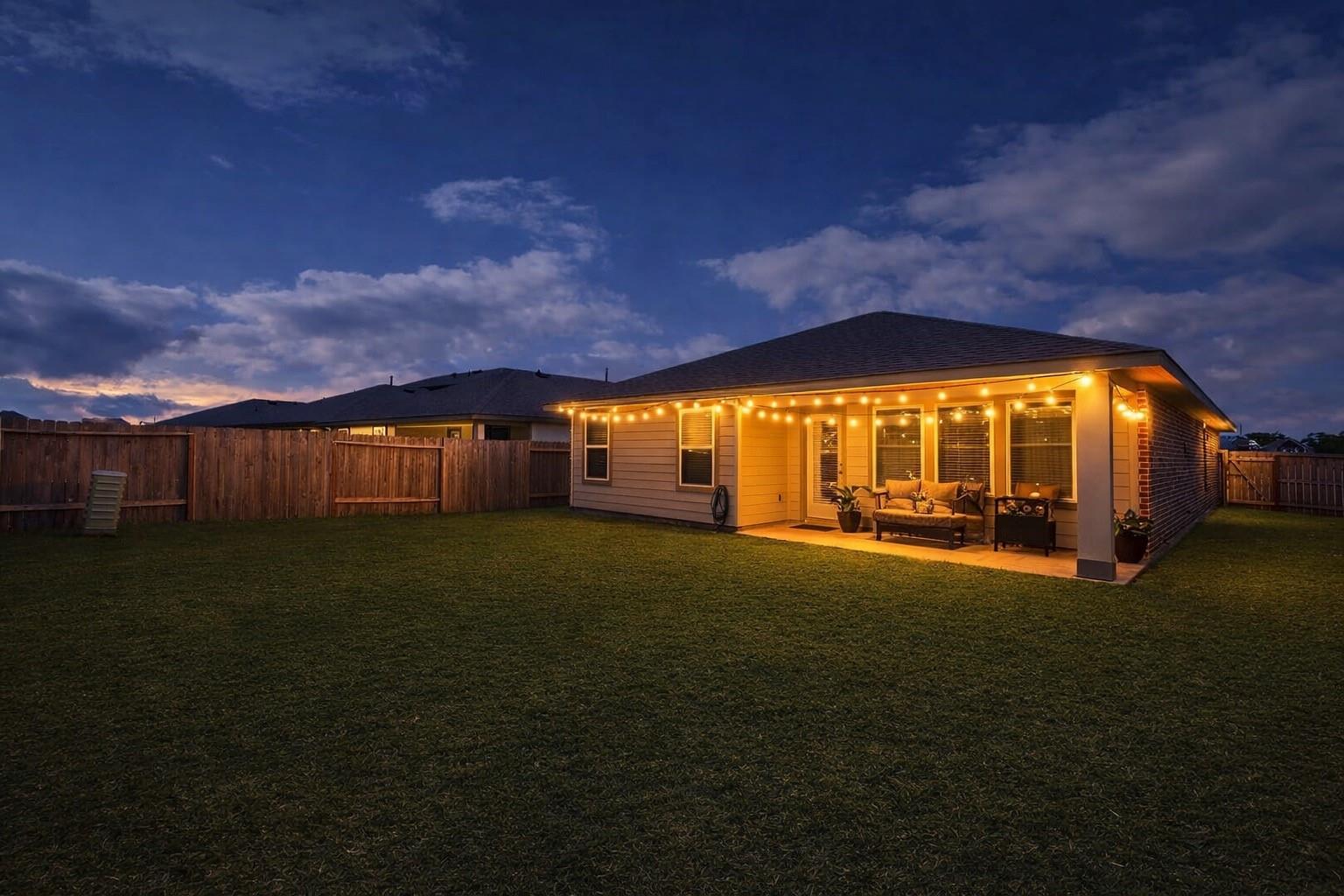 Home’s rear patio glowing with string lights at dusk, overlooking a wide fenced lawn.