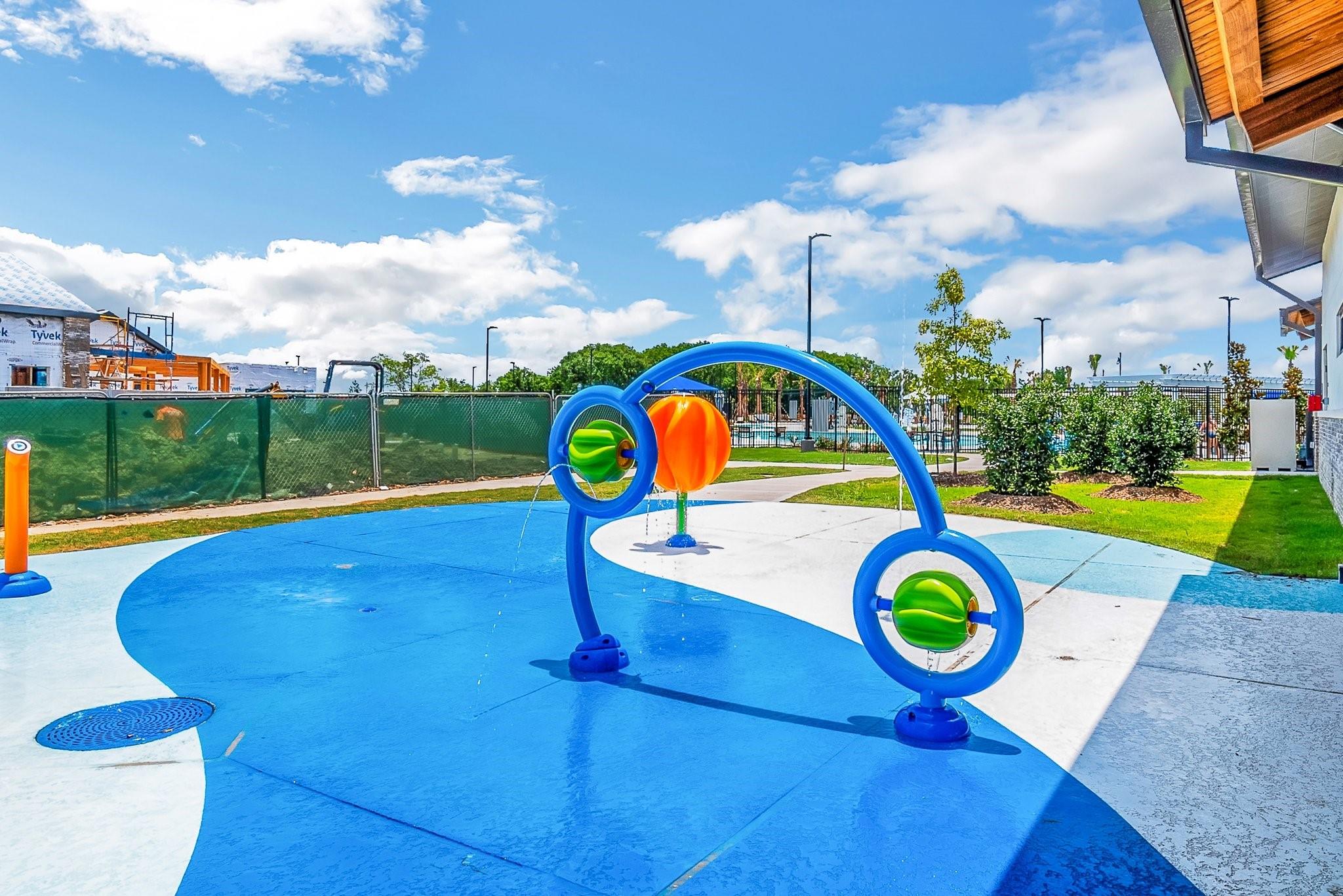 Community splash pad with colorful water-play equipment, blue surfacing, and a fenced pool in the background.
