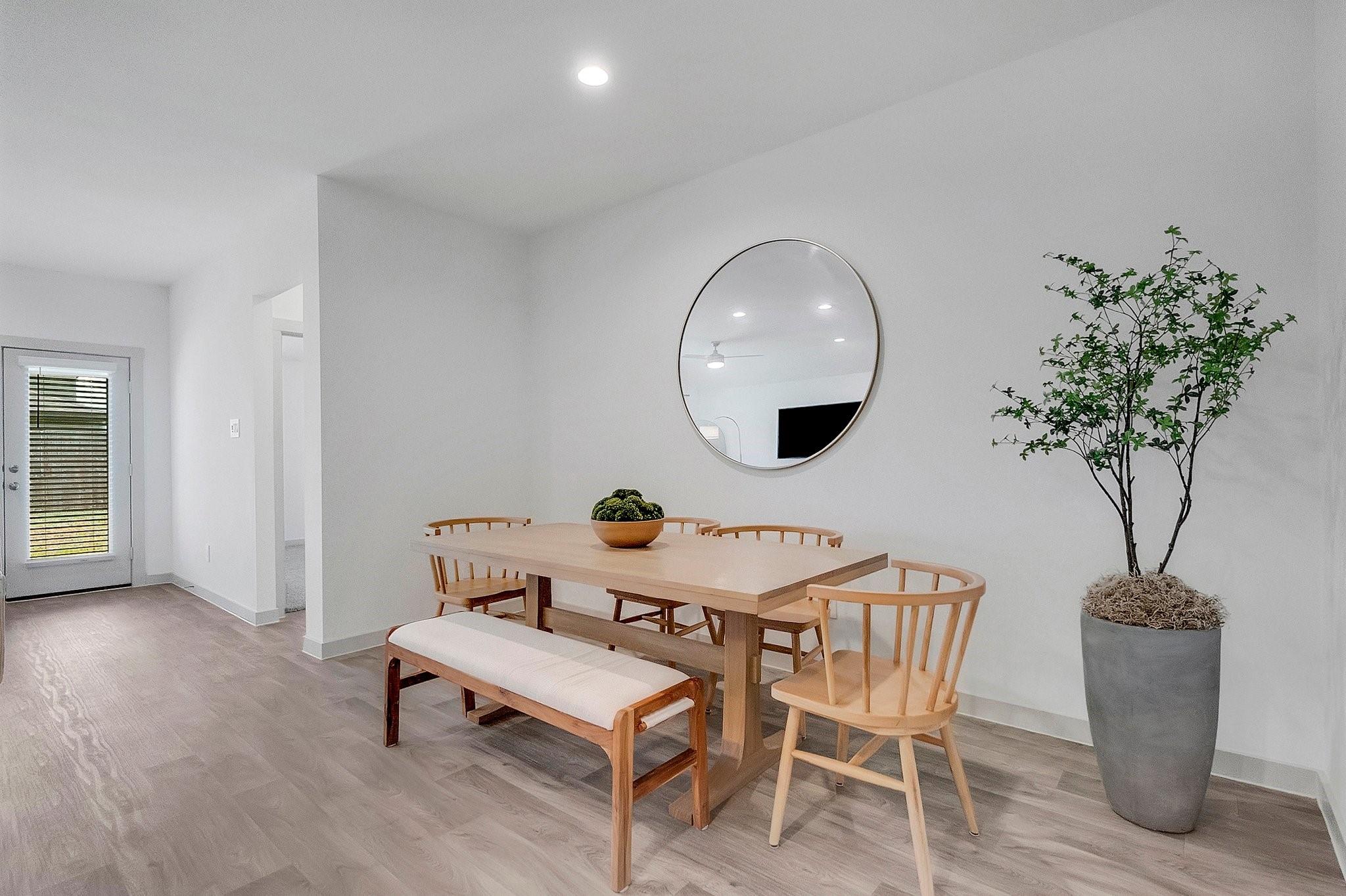 Dining area with a light wood table, bench, round mirror, and potted tree.