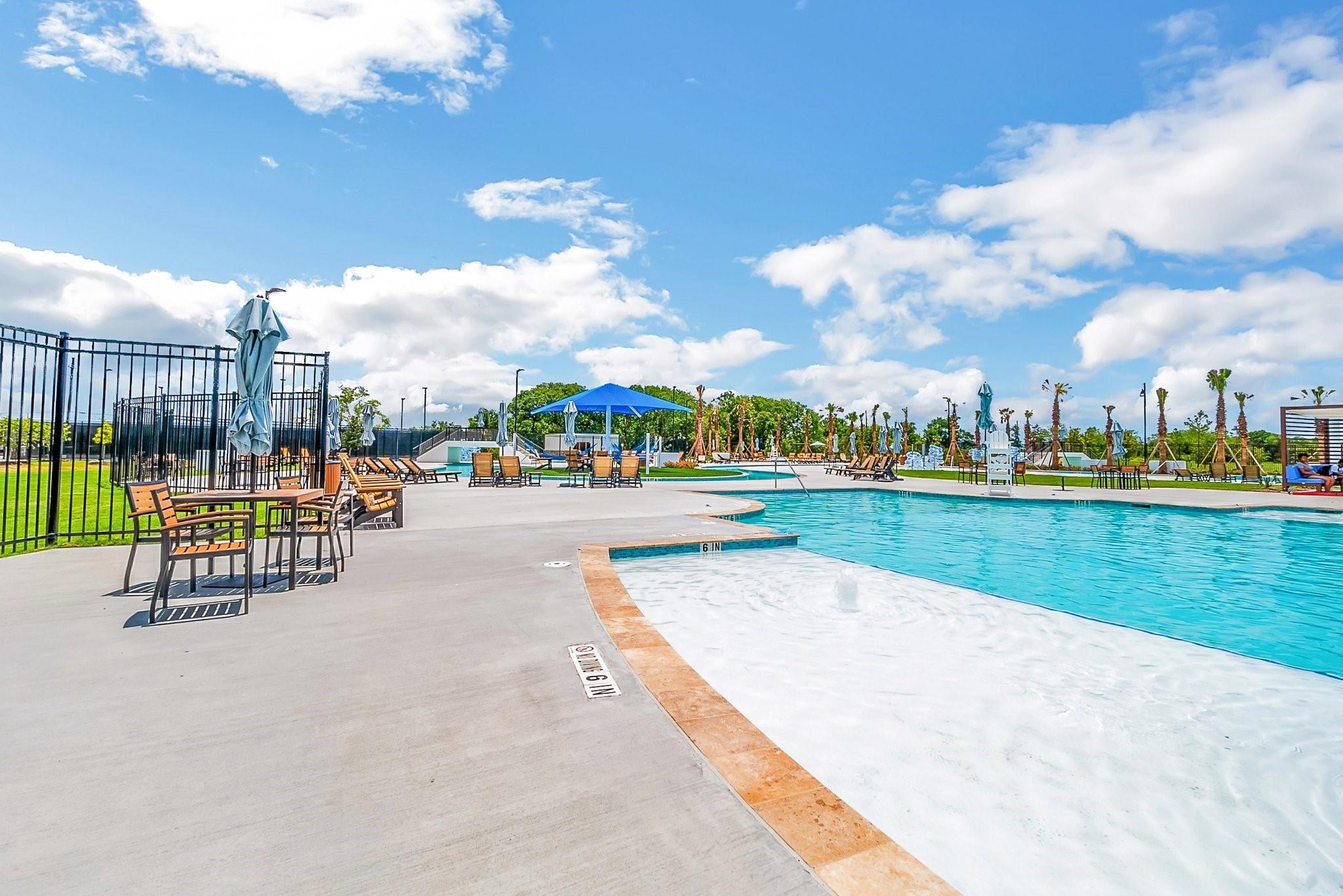 Outdoor community pool with a shallow entry, blue umbrella, lounge chairs, and palm trees.