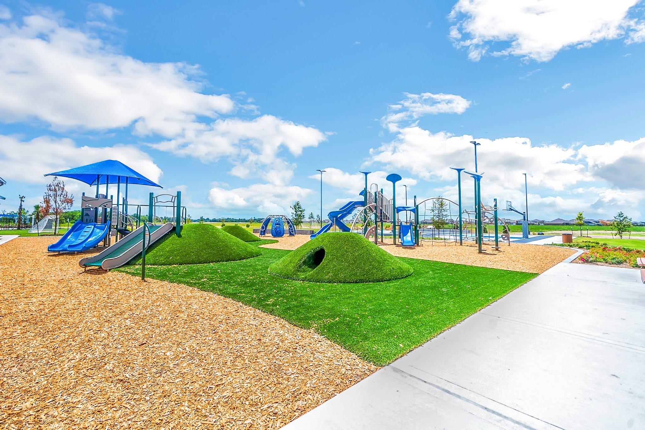 Community playground with blue slides, climbing structures, and artificial-grass mounds with a tunnel.