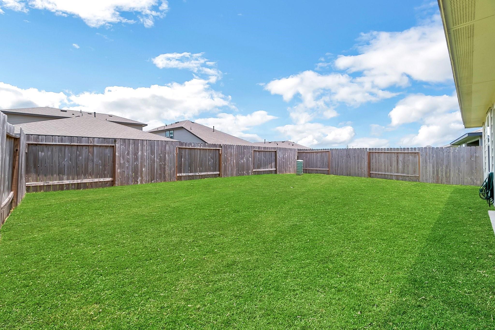 Fenced backyard with a wide green lawn and neighboring rooftops beyond the fence.