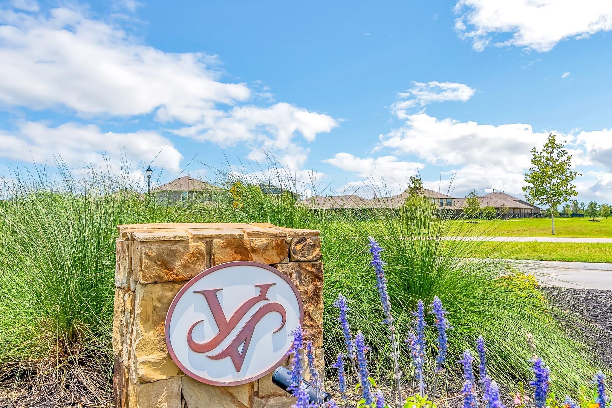 Stone neighborhood sign amid ornamental grasses, with homes and a green lawn behind it.