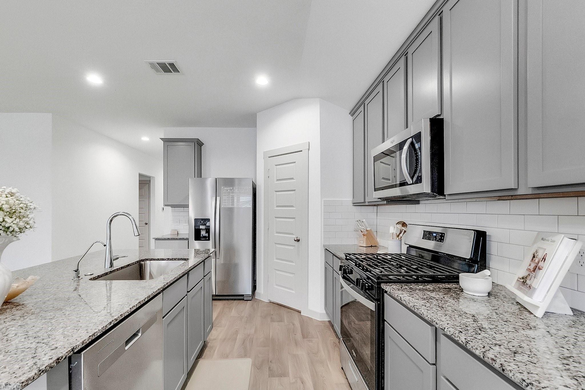 Modern kitchen with gray cabinets, granite counters, a large island sink, and stainless steel appliances.