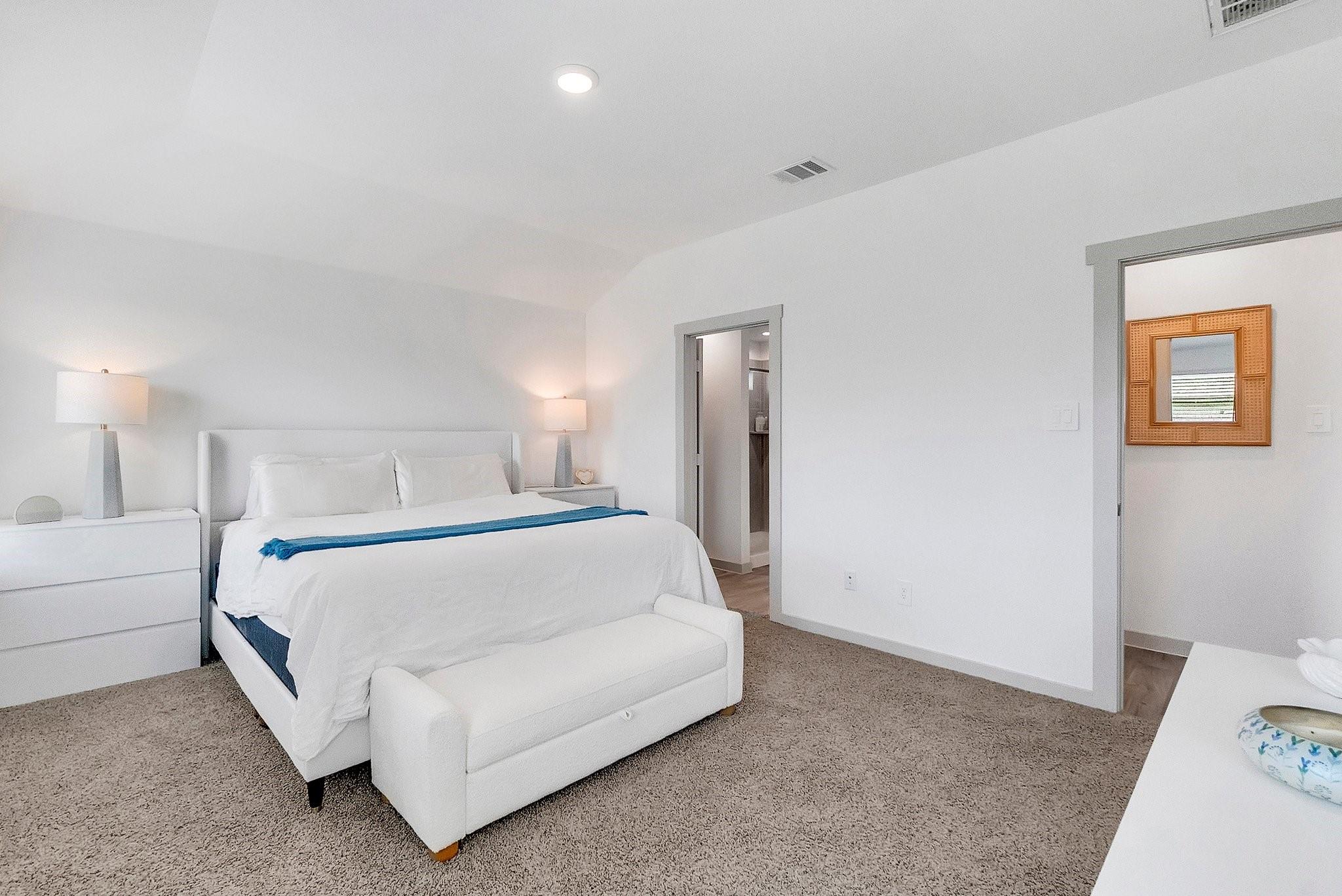 Bedroom with a white upholstered bed, blue throw, beige carpet, and doorway to an adjoining room.