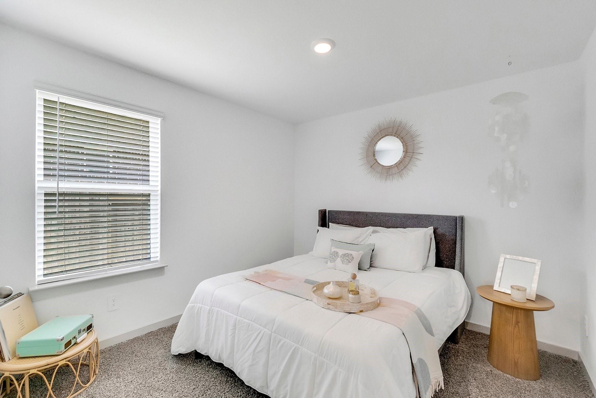 Staged bedroom with a white bed, sunburst mirror, and turquoise suitcase beside the window.