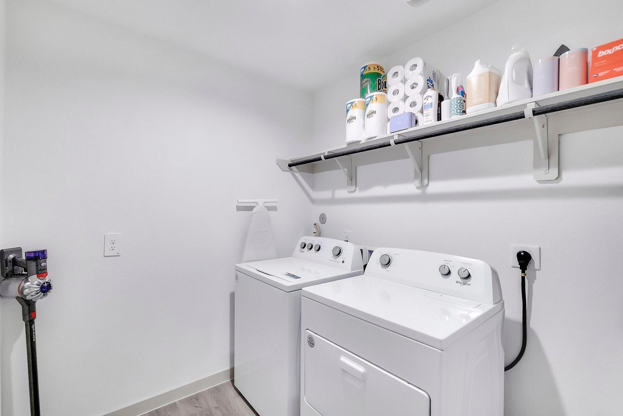 Laundry room with white washer and dryer beneath a long shelf of household supplies.