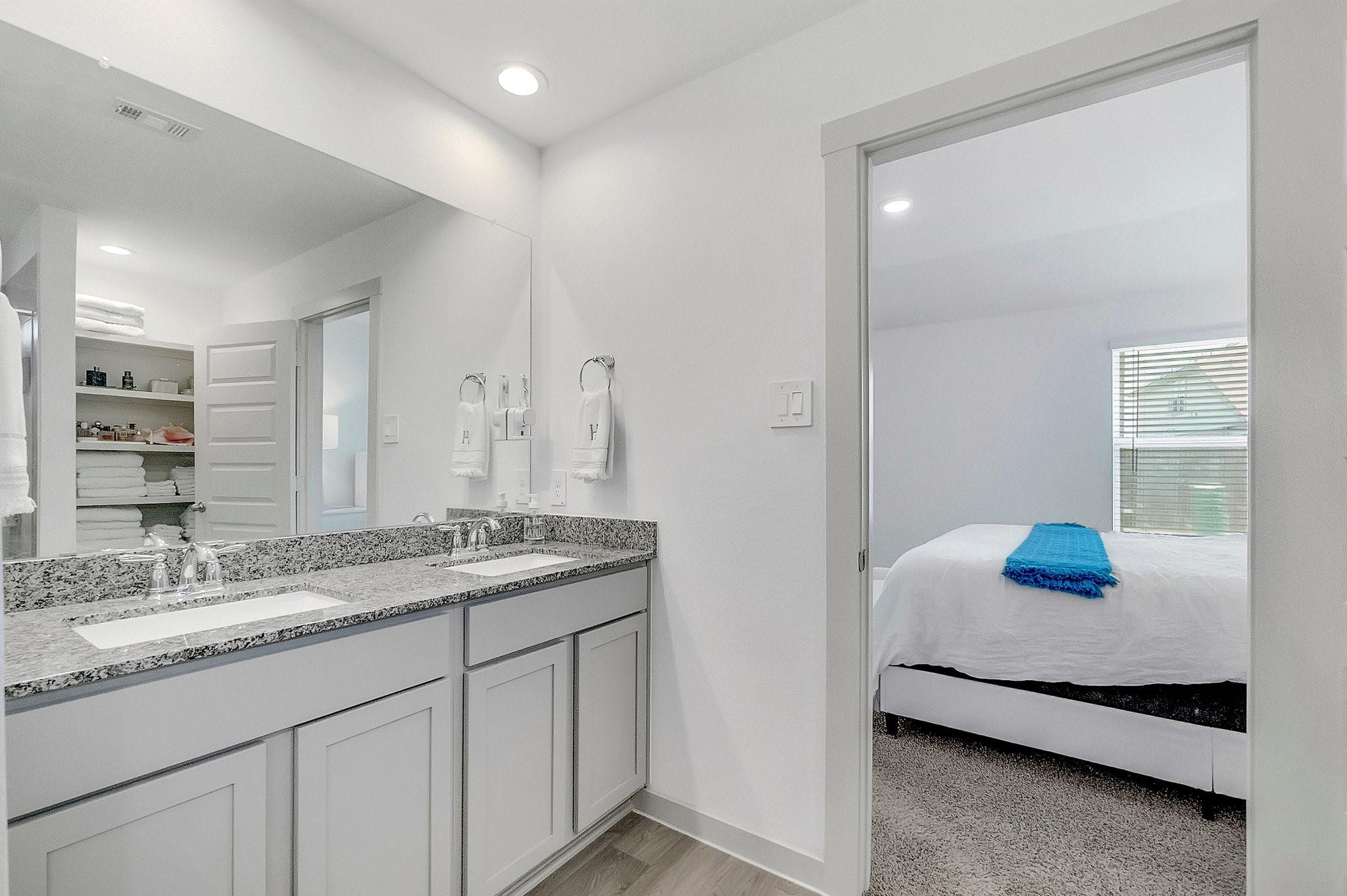 Bathroom with a double-sink granite vanity and mirror, opening onto a bedroom with a blue throw.